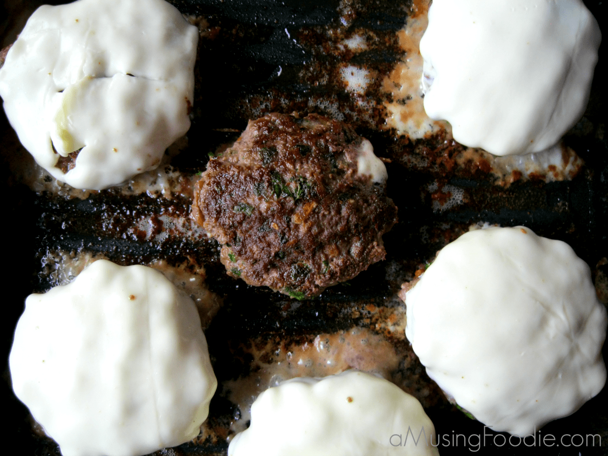 Slider patties cooking in a pan. Five of the six have melted white cheese on top.