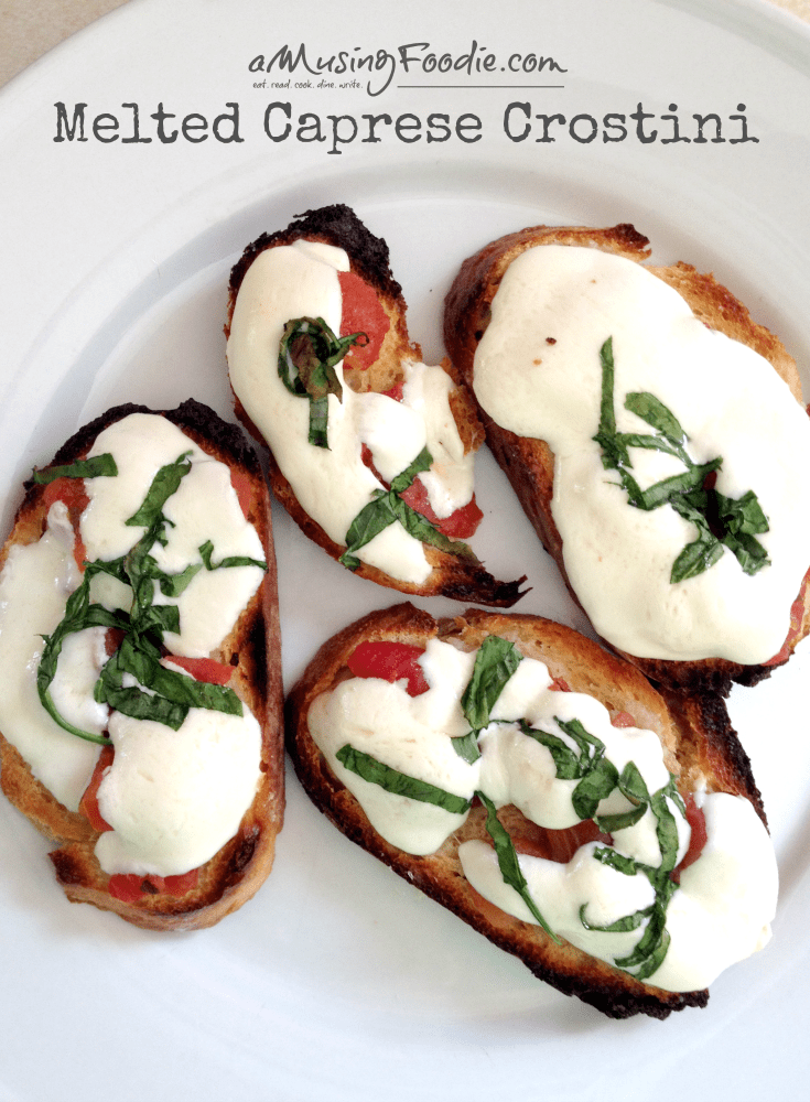 Melted caprese crostini with chopped fresh basil sitting on a white plate. The words "Melted Caprese Crostini" and "amusingfoodie.com" and "eat.read.cook.dine.write." are overlayed.