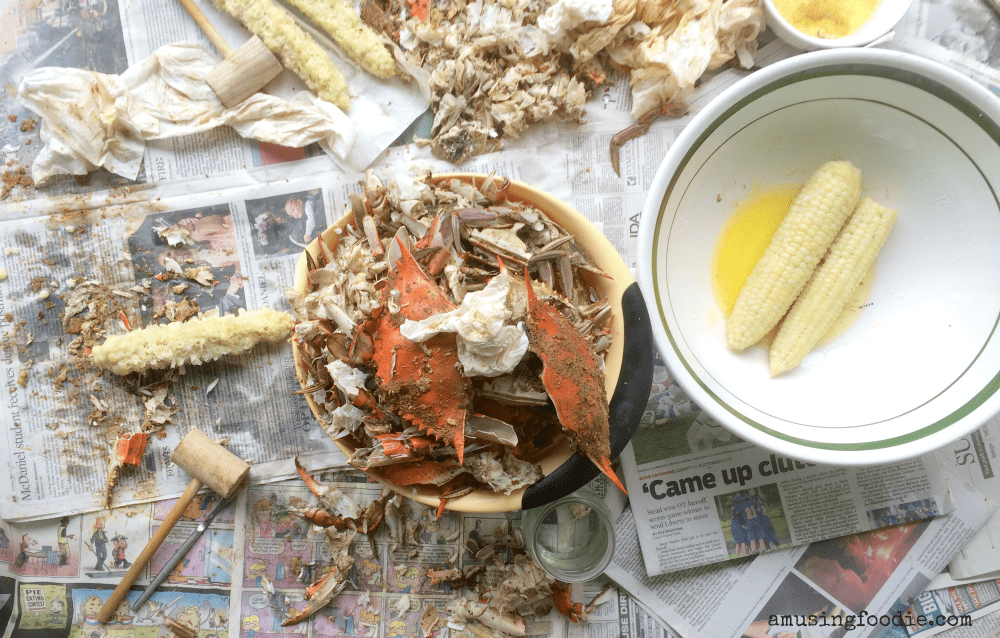 Overhead shot of empty blue crab shells in a yellow bowl on a table covered in newspaper. Eaten corn on the cob and crab mallets are on the table. Uneaten corn on the cob is in a bowl to the side.