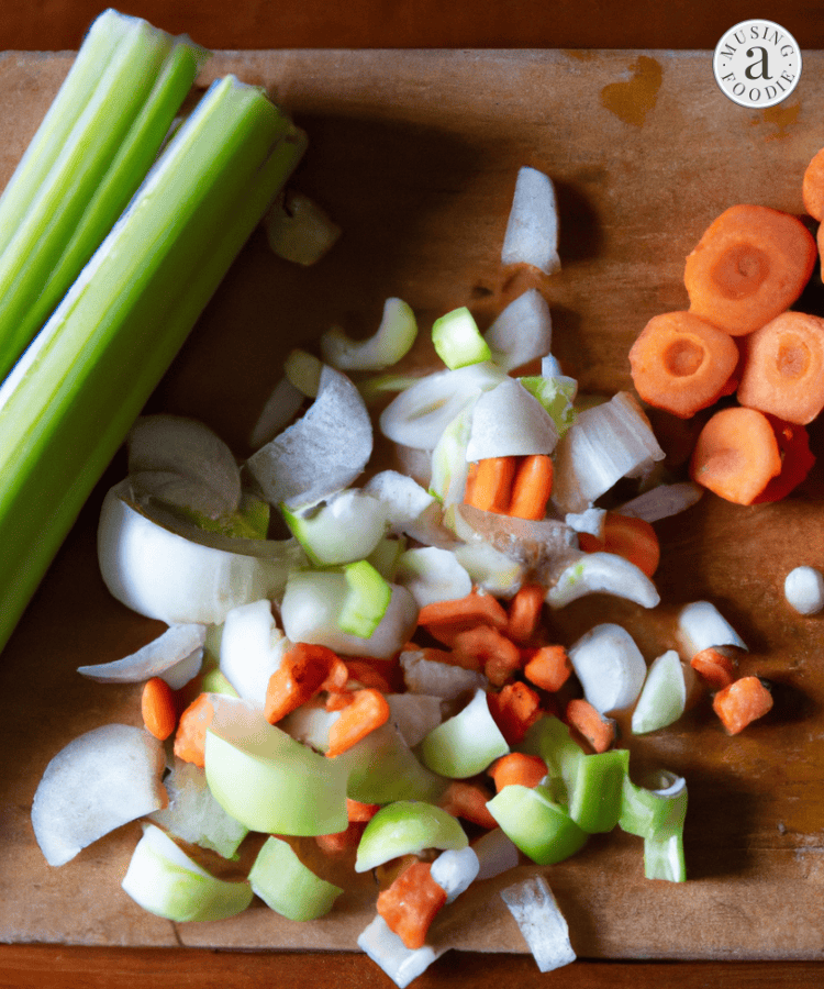 Photo in a kitchen with diffused natural window light of mis en place on a wooden cutting board, with diced carrots, onions, celery.